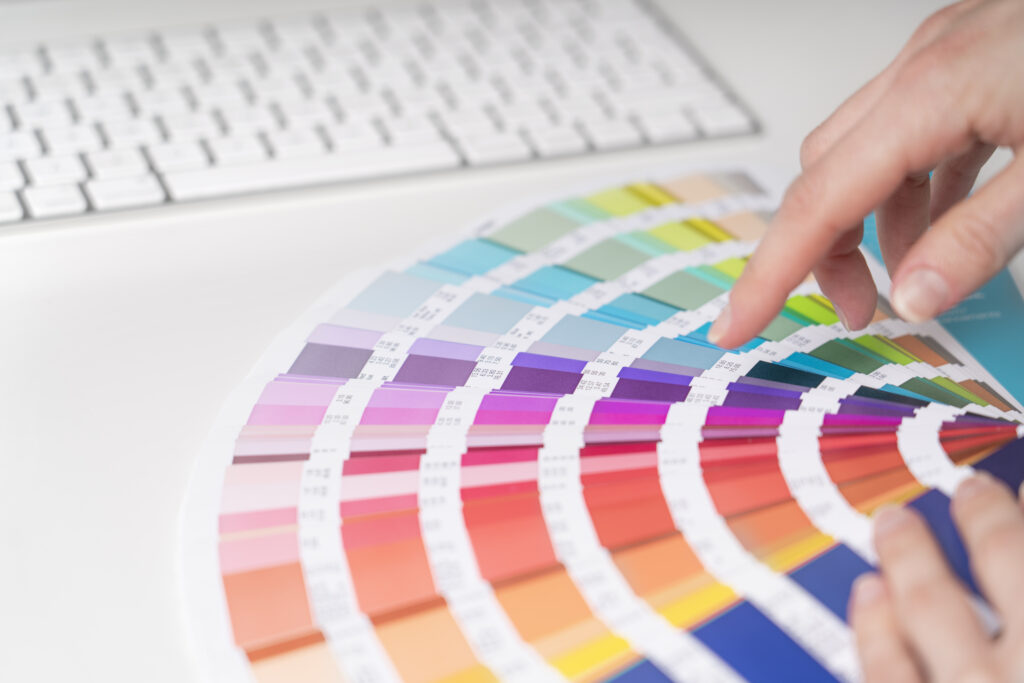 Hände blättern durch einen Farb-Fächer mit verschiedenen Pantone-Tönen vor einer Computertastatur.