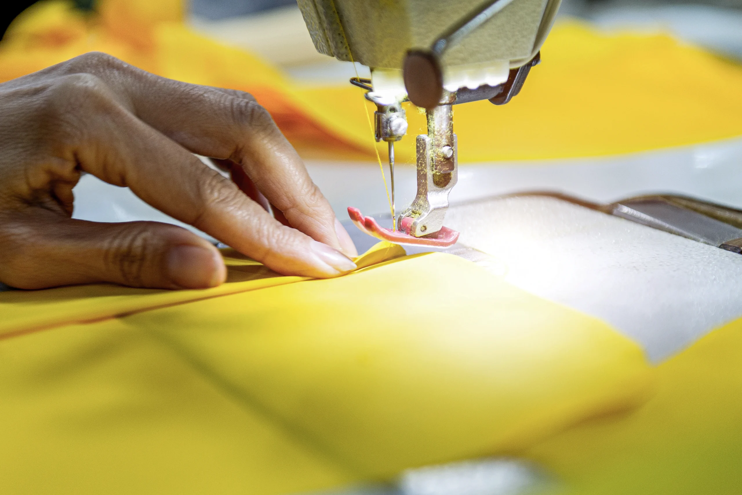Hand eines Arbeiters führt hellgelben Stoff unter der Nadel einer Industrienähmaschine.