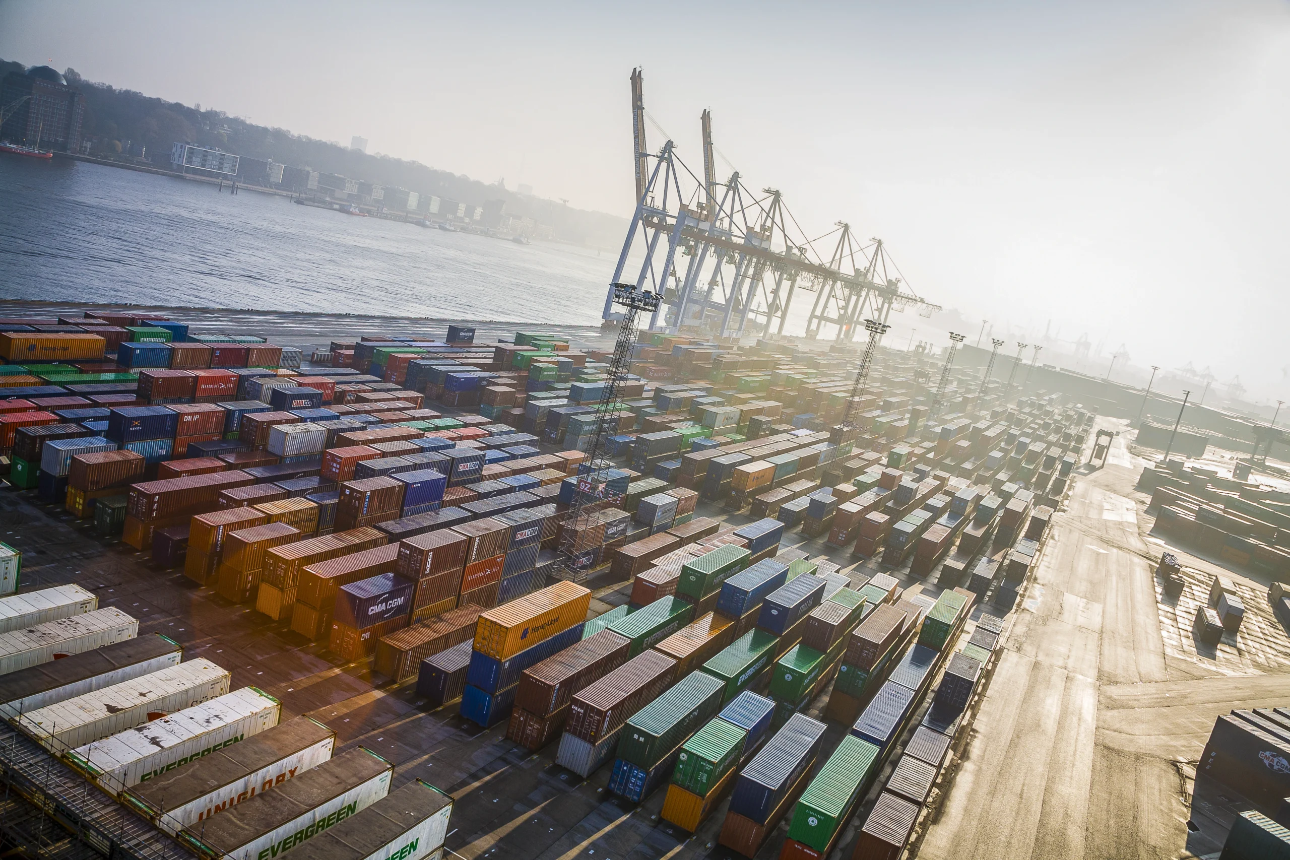 Luftaufnahme eines Containerhafens mit gestapelten Frachtcontainern und großen Verladekränen am Wasser.